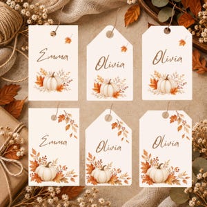 May include: Six rectangular and hexagonal paper gift tags featuring a white background, a central illustration of a white pumpkin surrounded by autumn leaves, and the printed names Emma or Olivia in a brown script font with twine loops.