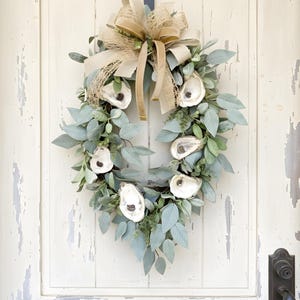 May include: A teardrop-shaped wreath featuring sage green eucalyptus leaves and five white oyster shells arranged throughout the foliage. A large, multi-layered bow made of tan burlap and beige velvet ribbon is secured at the top center.