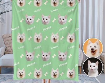 May include: A green fleece blanket with a repeating pattern of white dog and cat faces and bone shapes. The text "SIMBA" and "SOPHIA" are printed on the blanket. The blanket is being held by a person.