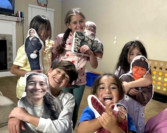 May include: A group of children smiling and holding custom-printed pillows featuring images of people. The pillows have various designs, including a person in a military uniform and another in camouflage. The children are in a living room setting.