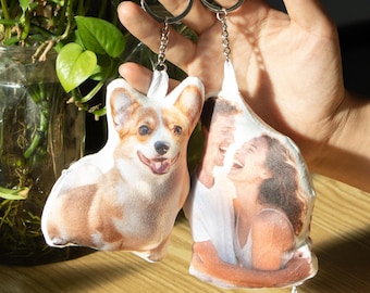 May include: Two custom keychains are held up in front of a green plant. One keychain is shaped like a Corgi dog with a smiling face, and the other keychain has a photo of a laughing couple. Both keychains have silver key rings.