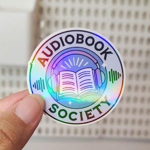 May include: A holographic sticker with a rainbow effect featuring a book with headphones and the text "Audiobook Society".