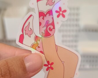Op de afbeelding: Een sticker met een paar roze en witte cowboylaarzen met bloemenmotieven. De laarzen bevinden zich op een paar benen met een roze bloem op de kuit.
