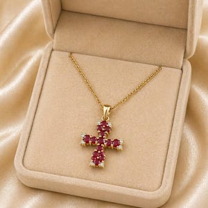 Può includere: Una collana color oro con un pendente a forma di croce. La croce è adornata con pietre preziose ovali rosso intenso e piccole pietre preziose trasparenti. La collana è presentata in un portagioie beige con fodera in raso.