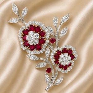 May include: A floral brooch featuring two flower blossoms with ruby and diamond accents. The center of each flower is white, surrounded by a ring of red rubies and a border of diamonds. The brooch has diamond-studded stems and leaves.