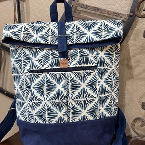 May include: A blue and white patterned backpack with a roll-top closure and a navy blue bottom. The backpack has a zippered pocket on the front and a leather tag that says "Handmade".