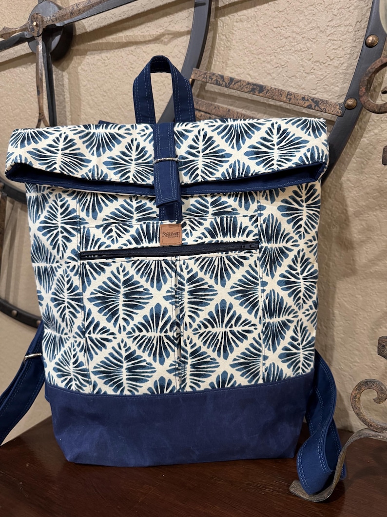 May include: A blue and white patterned backpack with a roll-top closure and a navy blue base. The backpack has a zipped pocket on the front and a leather tag that says "Handmade".