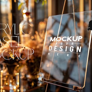Puede incluir: Una maqueta de caballete de acrílico transparente con un soporte dorado. El caballete está en blanco y listo para su diseño. El texto "MOCKUP YOUR DESIGN HERE" está impreso en el caballete. El fondo está desenfocado y muestra un arreglo floral.