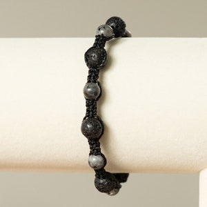 May include: A black beaded bracelet with alternating black lava rock and grey gemstone beads. The bracelet is made with black cord and is tied with a knot.