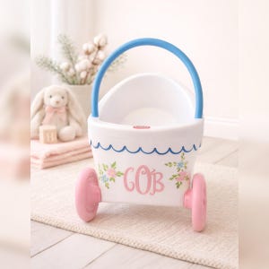 May include: A white and blue potty training seat with pink wheels and a blue handle. The seat is decorated with floral accents and the initials "GOB" in pink. A plush bunny and a stack of pink towels are in the background.