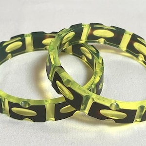 May include: Two circular bangles featuring a translucent neon yellow resin base with embedded dark brown, rectangular, and oval-shaped segments. The bracelets have a smooth, polished finish with a repeating pattern of alternating colors.