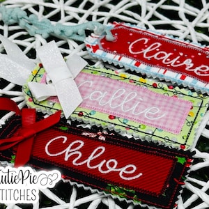 May include: Embroidered fabric gift tags in red, green, and pink with the names "Claire", "Callie", and "Chloe" in white script. Each tag has a decorative border and is adorned with ribbons and bows. The tags are on a white woven background.