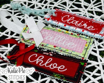 Pode incluir: Etiquetas de presente de tecido bordado em vermelho, verde e rosa com os nomes "Claire", "Callie" e "Chloe" em escrita branca. Cada etiqueta tem uma borda decorativa e é adornada com fitas e laços. As etiquetas estão em um fundo branco tecido.