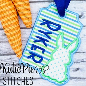 May include: A blue and green striped fabric tag with the name "Ryker" embroidered in blue. A white bunny with blue polka dots is embroidered on the tag. The tag is attached to a blue ribbon.