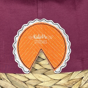 May include: A close-up of a burgundy fabric with a white and orange embroidered pie design. The pie is missing a slice and has a scalloped crust.