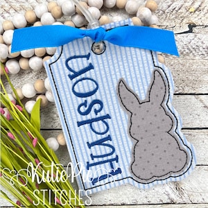 May include: A blue and white striped tag with the name "Hudson" in blue lettering. A gray bunny with polka dots is on the tag. A blue ribbon is tied at the top. The tag is part of a spring or Easter-themed craft.