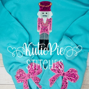 May include: Turquoise sweatshirt with a decorative nutcracker design. The nutcracker is wearing a pink jacket and a purple hat. Two pink sequined bows are on the lower part of the sweatshirt. The text "Kutie Pie Stitches" is also visible.