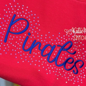 Puede incluir: Sudadera roja con la palabra "Pirates" bordada en azul, rodeada de pedrería iridiscente dispersa. El texto "Kutie Pie Stitches" está en escritura blanca en la esquina superior derecha.