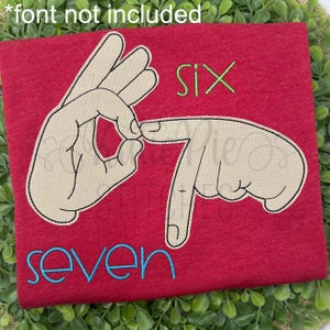 Puede incluir: Camiseta roja con un diseño que muestra los números seis y siete en Lengua de Signos Española. La palabra "seis" es verde y "siete" es azul.