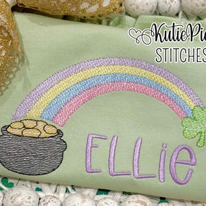 May include: A light green shirt with a rainbow, pot of gold, and shamrock embroidery. The name "Ellie" is embroidered in lavender script. The text "Kutie Pie Stitches" is at the top right.