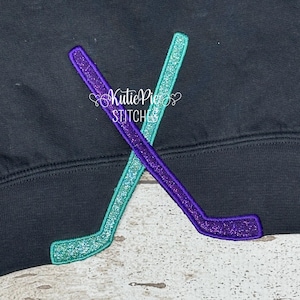 May include: Black sweatshirt with two crossed hockey sticks. One stick is purple, the other is teal, both with glittery accents. The text "Kutie Pies Stitches" is in white script.