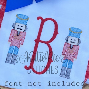 May include: White fabric with a red embroidered letter "B" and two embroidered nutcracker figures. The nutcrackers wear blue hats and red jackets. The text "Katie Pie STITCHES" is also embroidered on the fabric. A blue bow and red and white checkered fabric are in the background.