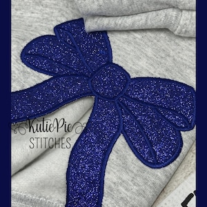 May include: Close-up of a gray sweatshirt with a large, sparkly blue bow applique. The bow is centered and has a glittery texture. The text "Kutie Pie Stitches" is printed in white below the bow.