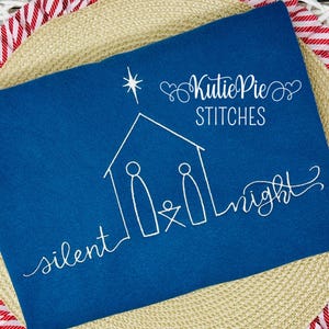 May include: A blue fabric with white embroidery of a nativity scene, a star, and the text "silent night". The text "Kutie Pie Stitches" is also embroidered on the fabric.