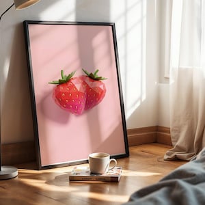 May include: A framed art print featuring two stylized strawberries in shades of red and pink against a light pink background. The print is in a black frame, and a coffee cup and book are on the floor.
