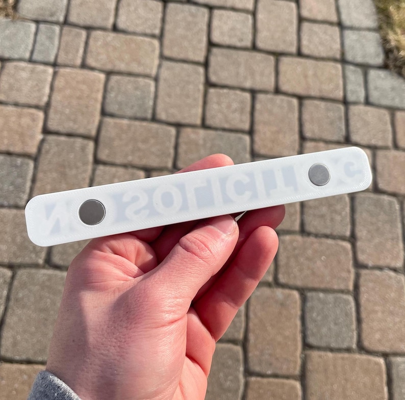 May include: White plastic magnetic strip with the text "ZOTICILOS" printed in black. The strip has two silver magnets on either end.
