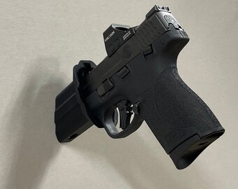 May include: A black handgun with a red dot sight attached to the top. The gun is mounted on a wall with a black holster.