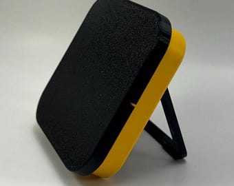 May include: A black and yellow stand-up device with a textured black surface and a yellow frame. The device has a black stand on the back, allowing it to stand upright. The design is simple and modern.