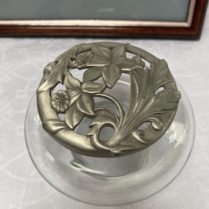 Könnte beinhalten: Ein silberfarbener Blumentopfdeckel aus Metall mit floralem Design. Der Deckel ist aus Zinn und zeigt ein zartes Blumenmuster mit Blättern und Blüten. Der Deckel ist so konzipiert, dass er auf einen gläsernen Blumentopf passt.