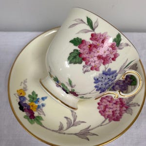Può includere: Servizio da tè color crema con design floreale di ortensie rosa, viola e blu. La tazza e il piattino hanno finiture dorate. La tazza da tè è inclinata sul piattino.