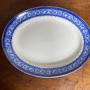May include: Oval serving platter with a white centre and a blue decorative border. The border features a repeating scroll design and a scalloped edge. The platter is made of ceramic and is suitable for serving food.