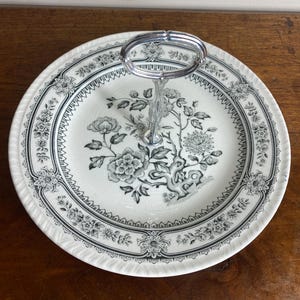May include: A two-tiered serving plate with a floral design in black and grey. The plate has a silver handle. The floral pattern includes flowers and leaves. The plate is on a wooden surface.