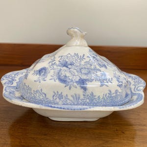 May include: A white ceramic butter dish with a blue floral pattern. The dish features a square base with scalloped edges and a matching lid with a decorative handle. The blue print depicts intricate flowers and foliage across the entire surface.