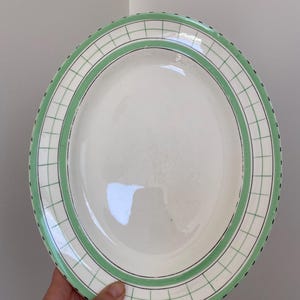 May include: An oval-shaped ceramic serving platter featuring a cream-coloured centre and a wide border decorated with a green grid pattern. The rim is accented with a thin black line and a series of small black dots along the outer edge.