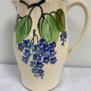 Puede incluir: Jarra de cerámica color crema con borde y asa negros. La jarra está decorada con hojas verdes, tallos negros y racimos de flores azules con centros amarillos. Una pieza clásica para servir bebidas.