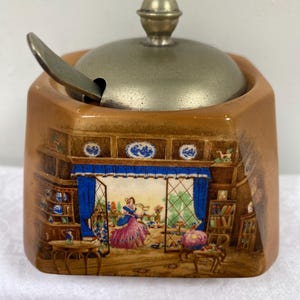 Könnte beinhalten: Eine Vintage-Zuckerdose mit einer detaillierten Innenszene mit einer Frau in einem rosa Kleid. Die braune Keramikschale hat einen Metalldeckel und einen Löffel. Die Szene umfasst blaue Vorhänge und Möbel.