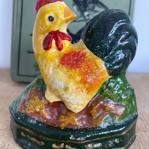 Puede incluir: Figura de gallo vintage pintada a mano. El gallo tiene cuerpo amarillo, cresta y barbilla rojas y cola negra. Está posado sobre una base verde y marrón con ribete dorado. La figura es probablemente de cerámica.