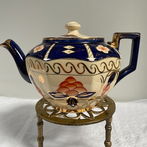 May include: A vintage teapot with a cream-colored base and navy blue accents. The teapot features gold trim and floral designs in red and orange. It sits on a decorative brass stand, showcasing a classic, elegant design.