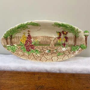 May include: Oval ceramic serving platter with a garden scene. The platter features a raised relief design of a garden with flowers, a fence, and trees. The colors include green, yellow, red, and brown. The platter is approximately 40 cm long.