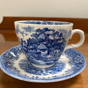 Könnte beinhalten: Blau-weißes Keramik-Tassen- und Untertassen-Set. Die Tasse und die Untertasse zeigen eine detaillierte Landschaftsszene mit Gebäuden und Bäumen. Das Design ist ein klassisches Toile-Muster mit weißem Hintergrund und blauen Illustrationen.