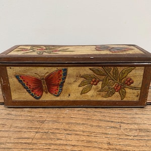 May include: Rectangular wooden box with a hinged lid. The box is painted with a butterfly and floral design in red, blue, and green on a cream background. The box has a dark brown frame.