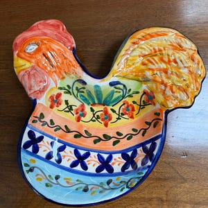 May include: A brightly coloured ceramic serving dish in the shape of a rooster. It has a red comb, orange head, and yellow and orange tail feathers. The body is decorated with floral patterns in orange, blue, and yellow, with a blue border.
