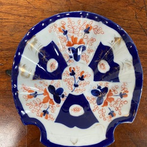 May include: A small, shell-shaped dish with a white interior and a blue and orange floral design. The rim is blue with gold accents. The dish is likely made of porcelain or ceramic.