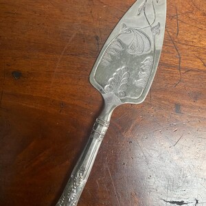 Puede incluir: Servidor de pastel plateado con diseños florales y de hojas ornamentados. El servidor presenta una hoja ancha y triangular con patrones botánicos grabados y un mango detallado. El mango tiene un diseño decorativo y texturizado.