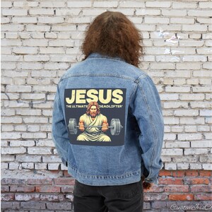 Puede incluir: Una chaqueta vaquera azul con un parche en la espalda que dice "Jesús, el levantador de peso muerto definitivo" y presenta una ilustración de Jesús levantando una barra.