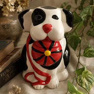 May include: A ceramic dog figurine featuring a white body with black patches, a red nose, and black ears. The dog holds a red and black ball with a yellow center in its mouth, attached to a red leash that drapes down the front of the figure.